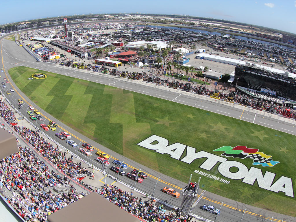 Vue aérienne du Daytona International Speedway pendant la course Daytona 500, avec des voitures de course sur la piste et une foule de spectateurs rassemblés dans les tribunes.