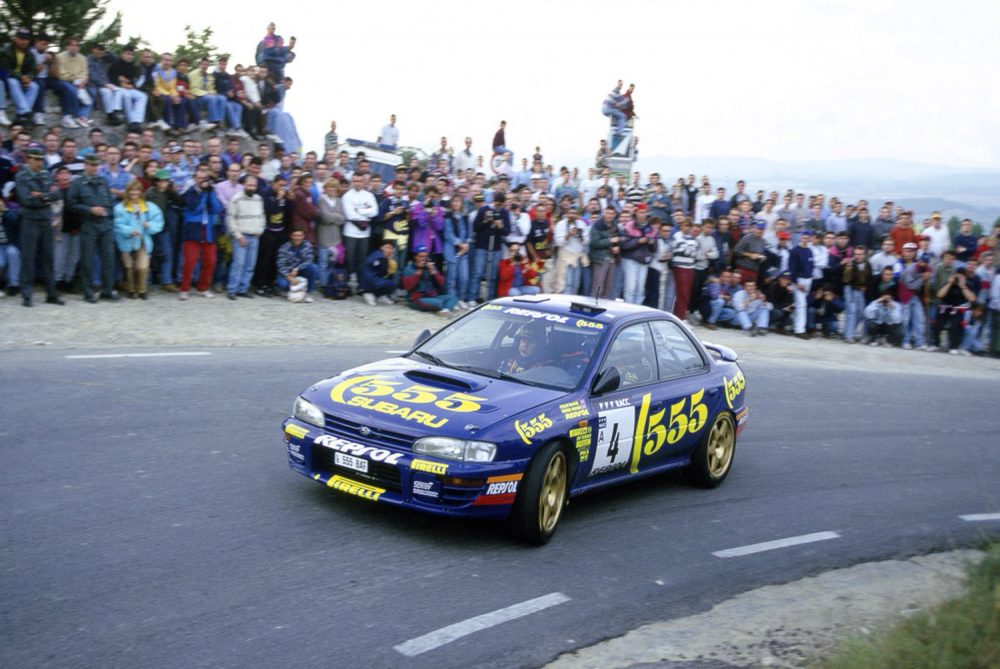 Une Subaru Impreza 555 avec le numéro 4, conduite par Carlos Sainz, prenant un virage pendant le Rallye Monte-Carlo 1995, avec une foule de spectateurs rassemblés sur le côté de la route.