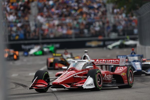 Une voiture de course rouge avec des sponsors, se disputant sur le circuit urbain de Détroit lors du Chevrolet Detroit Grand Prix, entourée de plusieurs autres voitures et d'une foule enthousiaste en arrière-plan.
