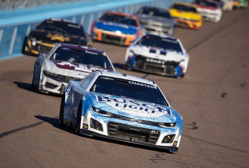 Voiture de course de Ross Chastain en tête, avec d'autres voitures de la NASCAR Cup Series en arrière-plan sur le circuit.
