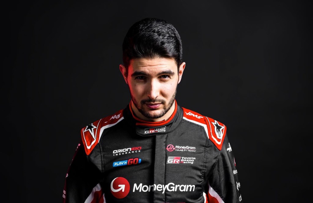 Un pilote de Formule 1, portant une combinaison de l'équipe Haas F1, se tient en portrait avec un regard sérieux sur fond noir.