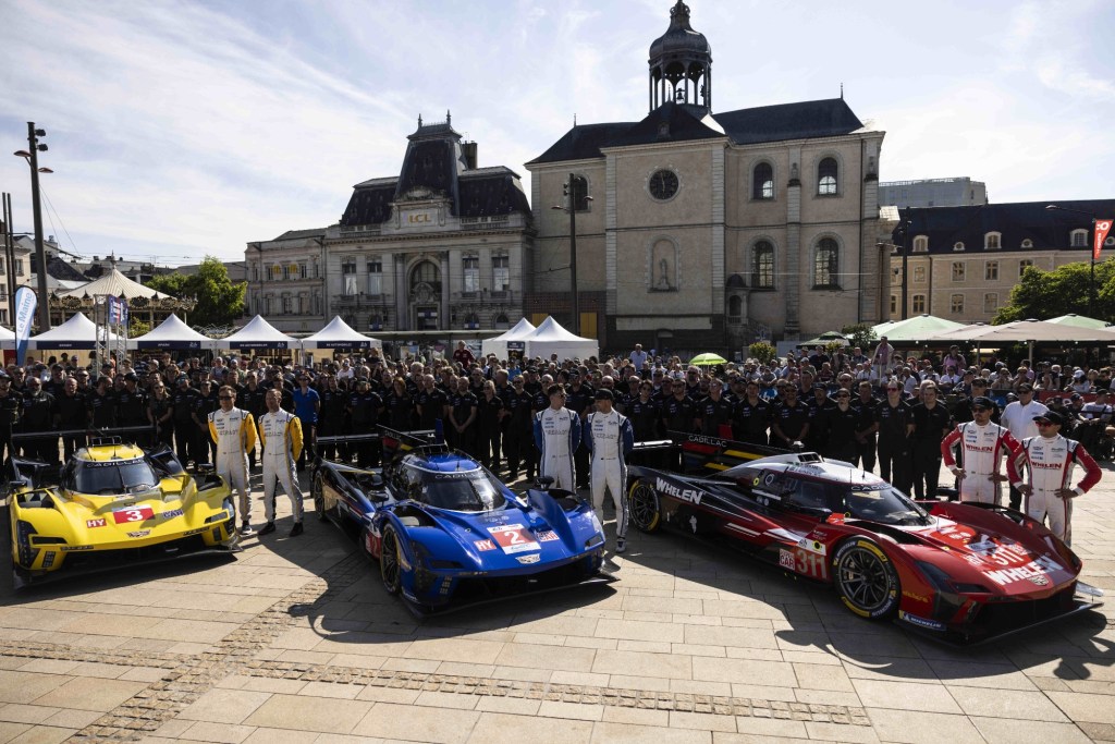 Des pilotes et équipes pose devant des voitures de course au centre-ville du Mans, avec des bâtiments historiques en arrière-plan.