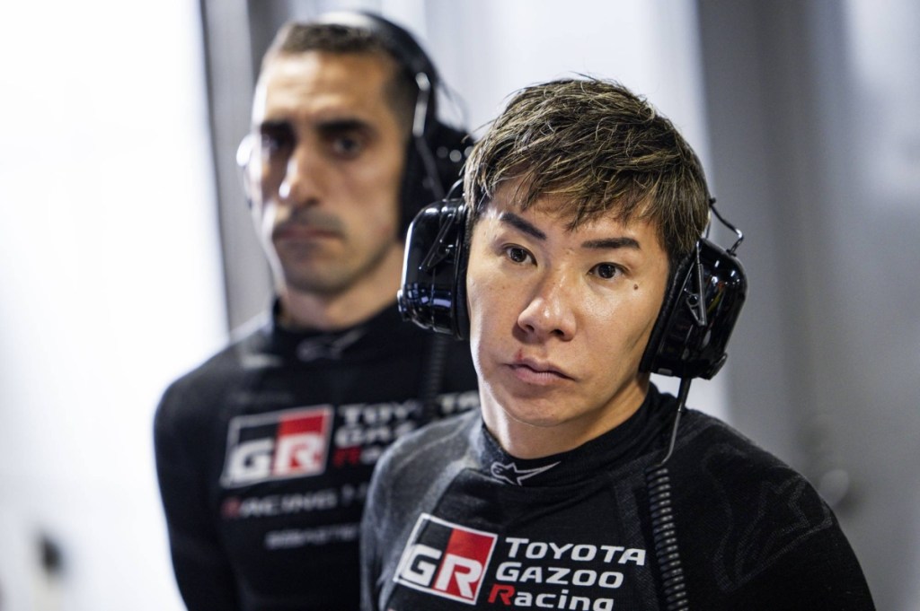 Kamui Kobayashi, pilote et Team Principal de Toyota Gazoo Racing, en tenue de course dans un garage, avec un coéquipier en arrière-plan.