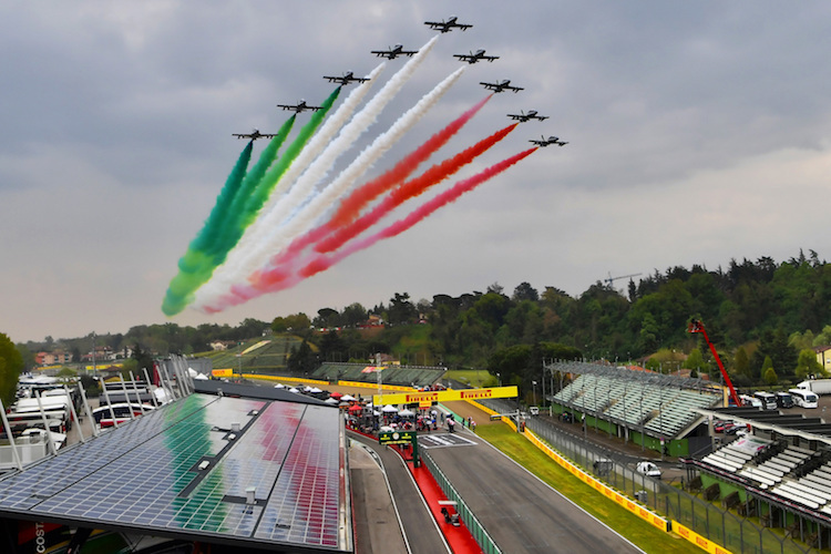 Avion de chasse en formation laissant des traînées de fumée tricolores sur le circuit d'Imola, avec des nuages en arrière-plan.