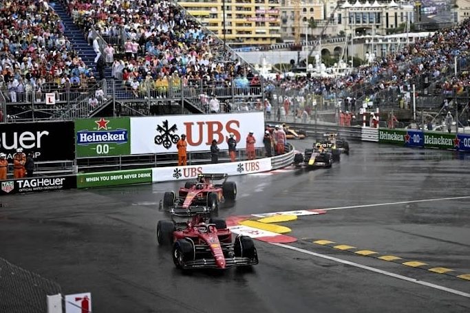 Course de Formule 1 sous la pluie, avec plusieurs voitures de course, dont des Ferrari, circulant sur le circuit de Monaco. Des spectateurs et des panneaux sponsors sont visibles en arrière-plan.