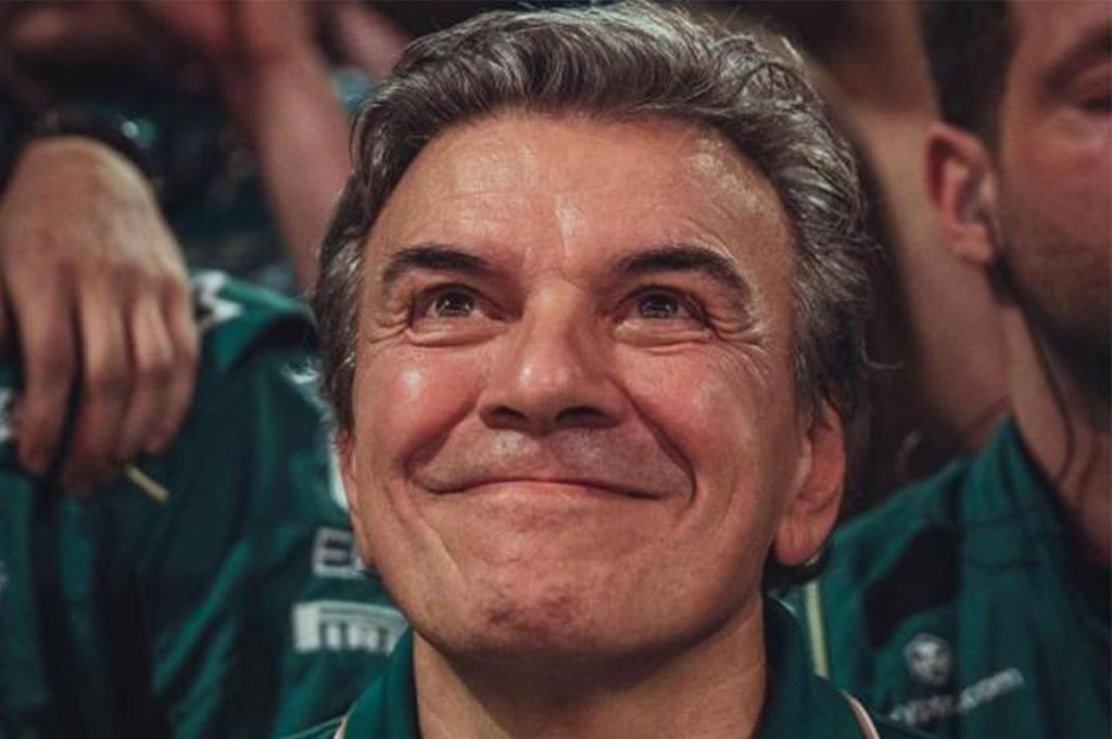 Un homme souriant, avec des cheveux grisonnants, entouré de personnes dans un environnement de sport automobile, portant une tenue verte.