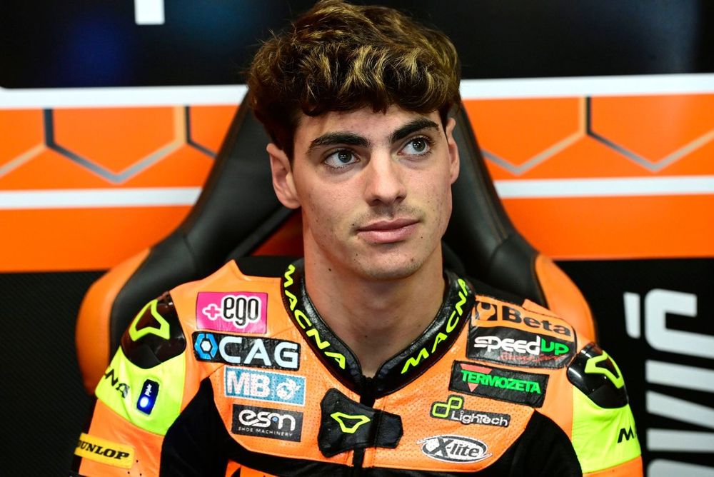 Un jeune pilote de moto avec des cheveux bruns assis, portant une combinaison de course colorée avec des logos d'équipe, dans un environnement de paddock.