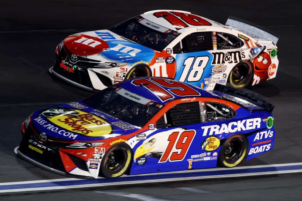 Deux voitures de course NASCAR en pleine compétition sur une piste, avec l'une portant les couleurs de M&M's et l'autre aux couleurs de Tracker.