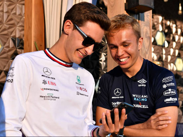 Deux pilotes de Formule 1 souriant et regardant un téléphone ensemble, vêtus de leurs uniformes de course.