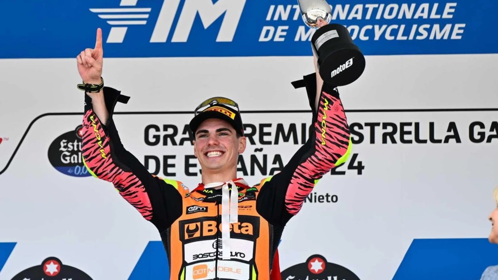 Un jeune pilote de MotoGP souriant sur le podium, levant un trophée avec une main, portant un design de combinaison de course coloré et une casquette, devant un panneau de course.