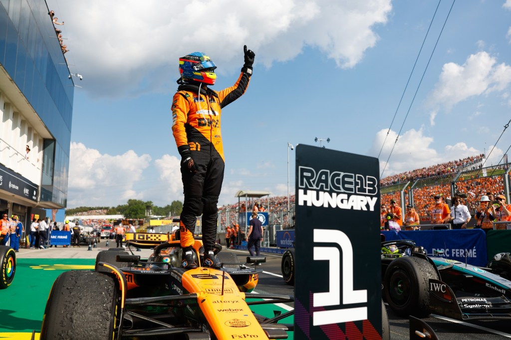 Pilote de Formule 1 célébrant sa victoire sur le podium à Budapest, brandissant le doigt en signe de numéro 1, entouré de spectateurs en arrière-plan.