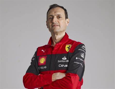 Un homme portant un uniforme de l'équipe Ferrari, avec les bras croisés et un léger sourire, se tenant debout en studio.