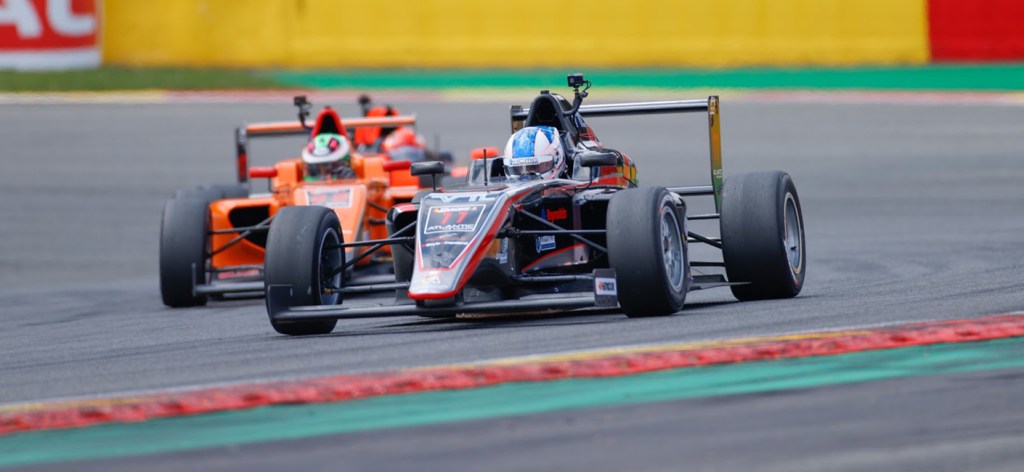 Une voiture de Formule 2 en course sur un circuit, avec un pilote en tête et une autre voiture en arrière-plan.