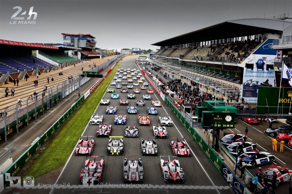 Vue aérienne de la piste des 24 Heures du Mans avec plusieurs voitures de course disposées en formation sur la grille de départ, entourées de spectateurs et des stands.