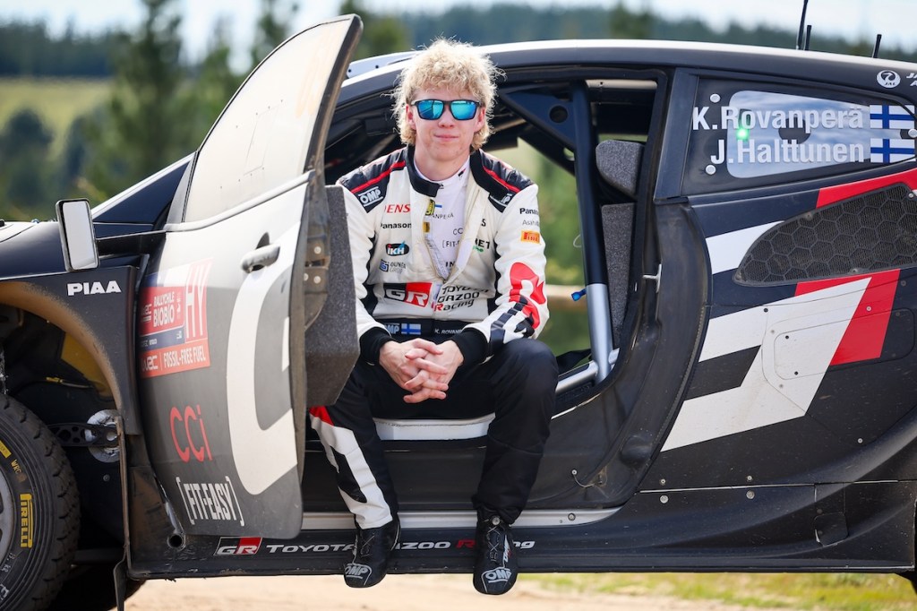Kalle Rovanperä assis sur le bord d'une voiture de rallye, souriant et portant une combinaison de course, avec des lunettes de soleil, dans un environnement extérieur.