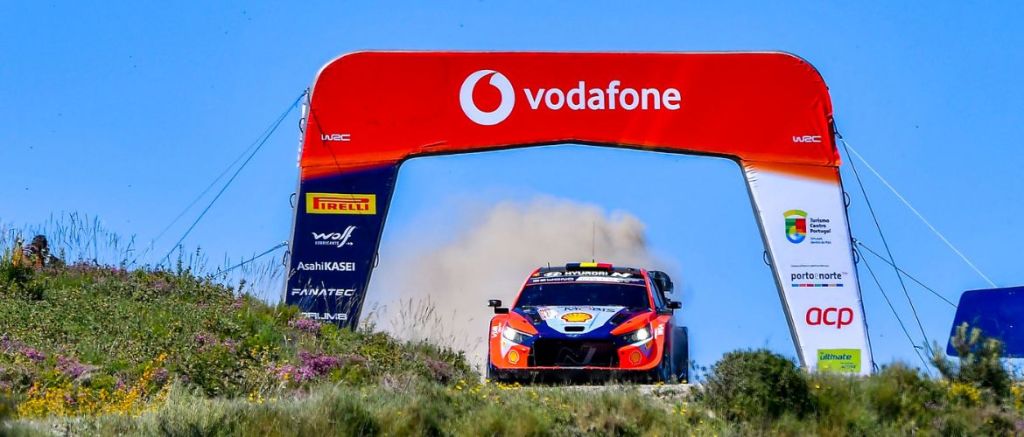 Une voiture de rallye passant sous un arc d'arrivée aux couleurs de Vodafone, avec de la poussière soulevée par le mouvement, dans un environnement naturel verdoyant.