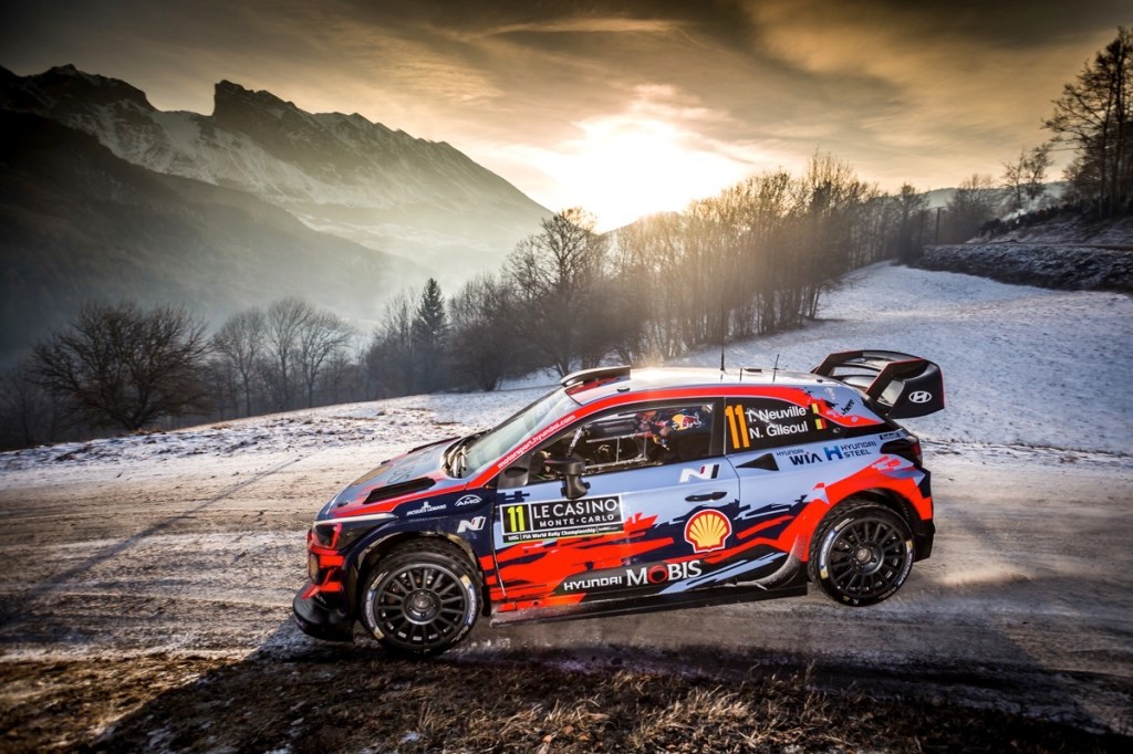 Voiture de rallye sur une route enneigée des Alpes, avec des montagnes en arrière-plan et un ciel partiellement nuageux.