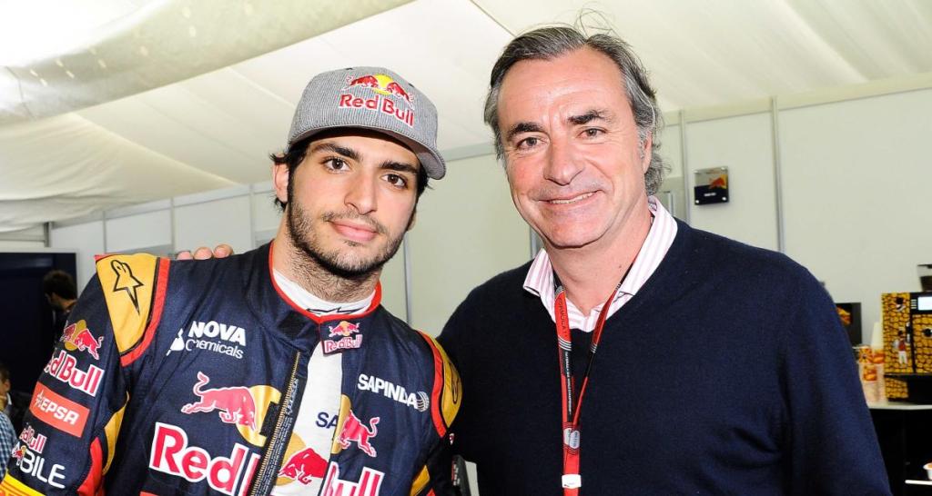 Carlos Sainz Jr. et Carlos Sainz Sr. posent ensemble, vêtus de vêtements associés à la course automobile, avec un fond flou d'événements sportifs.