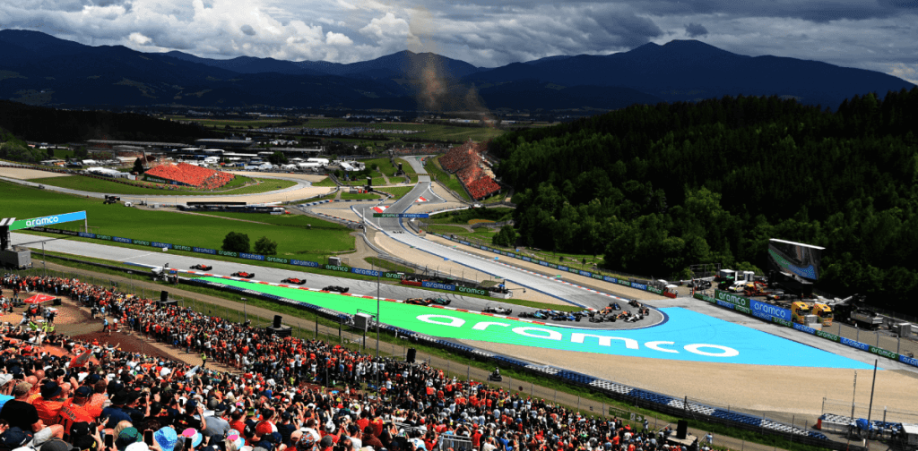Vue panoramique d'un circuit de course avec des voitures de course sur la piste, des spectateurs dans les tribunes, et des montagnes en arrière-plan.