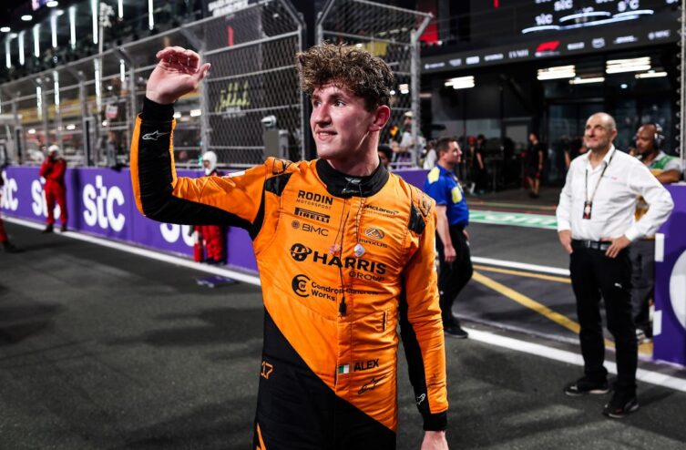 Alex Dunne, pilote de la McLaren Driver Academy, en tenue de course orange, lève la main pour saluer après une session sur le circuit, entouré de membres de l'équipe et de l'infrastructure de la course.