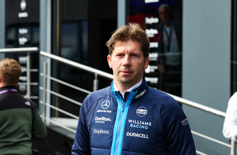 James Vowles, directeur d'écurie de Williams Racing, en tenue officielle, se tient devant un bâtiment de l'équipe pendant un événement de Formule 1.