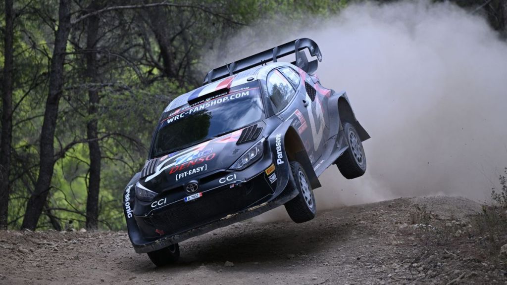Une voiture de rallye volant en l'air sur une piste de gravier, entourée d'arbres, capturant l'intensité du Rallye de l'Acropole.