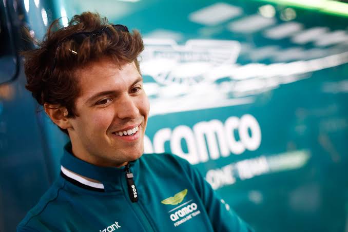 Felipe Drugovich, pilote brésilien et réserviste chez Aston Martin, souriant devant un mur de garage de l'équipe.
