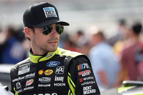Un pilote de NASCAR portant une casquette et des lunettes de soleil, avec une combinaison de course aux couleurs vives, se tenant près de sa voiture de course en arrière-plan.