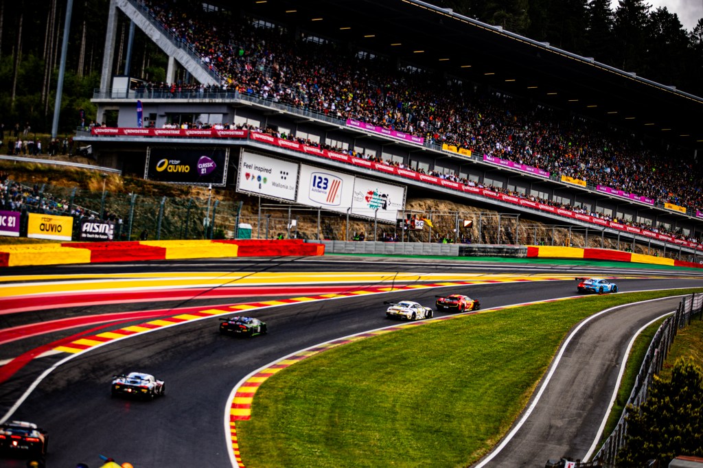 Vue panoramique du Circuit de Spa-Francorchamps pendant une course, avec des voitures de course sur la piste et une foule de spectateurs dans les tribunes.