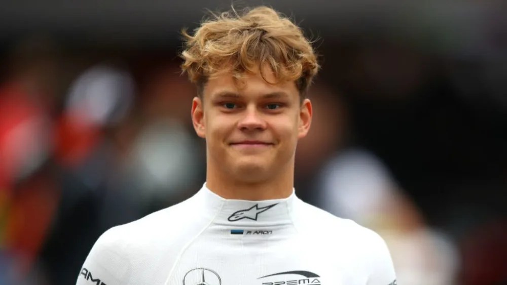 Portrait d'un jeune pilote en combinaison de course, sourire aux lèvres, avec une foule floue en arrière-plan.